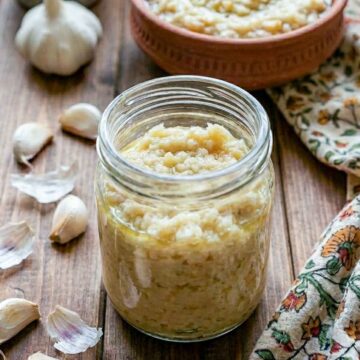 Jar of garlic paste with olive oil, surrounded by fresh garlic cloves on a wooden table.