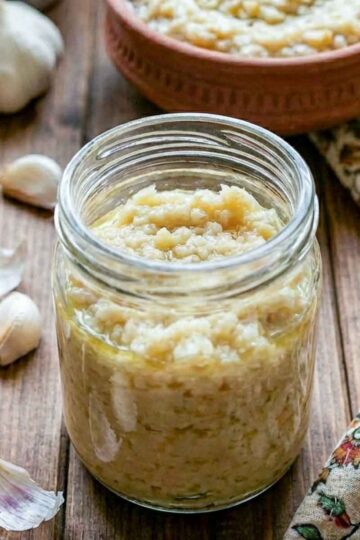 Jar of garlic paste with olive oil, surrounded by fresh garlic cloves on a wooden table.