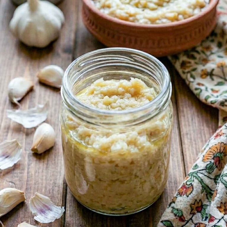 Jar of garlic paste with olive oil, surrounded by fresh garlic cloves on a wooden table.