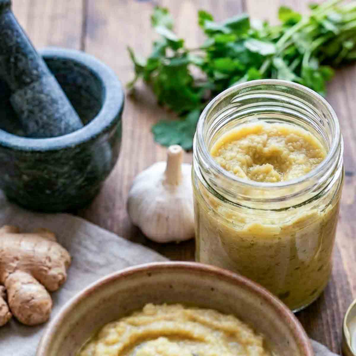 Jar of ginger garlic paste with fresh ginger, garlic, and mortar on a wooden table.
