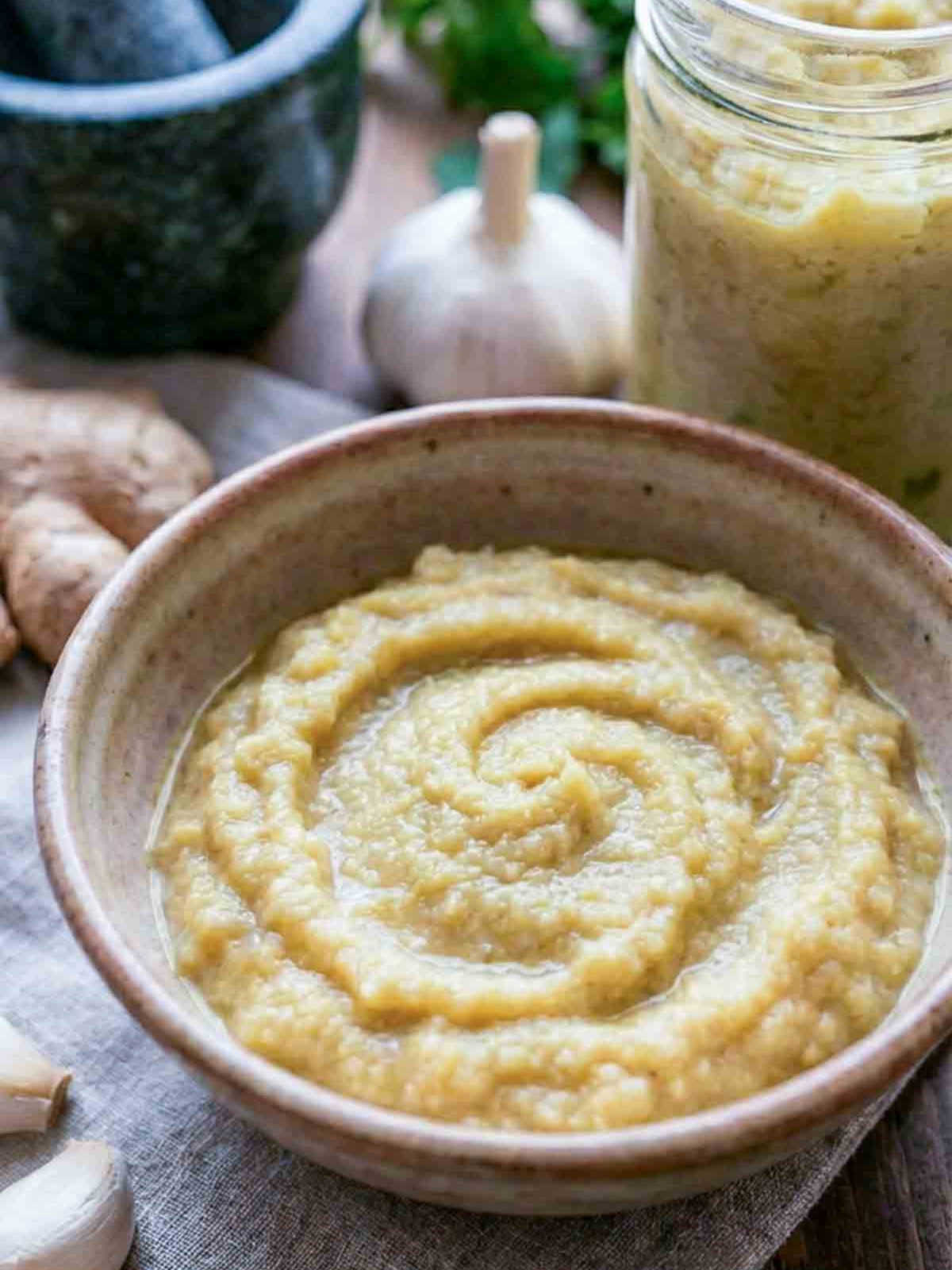 Bowl of ginger garlic paste with jar, fresh ginger, and garlic on a wooden table.
