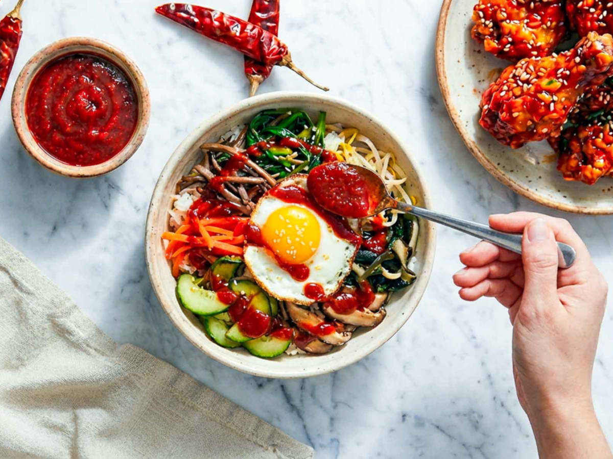 Hand spooning gochujang sauce over a bibimbap bowl with vegetables and fried egg.