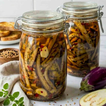 Close-up of two jars of pickled eggplant with garlic, herbs, and chili flakes, with fresh eggplant and seasonings arranged on a countertop.