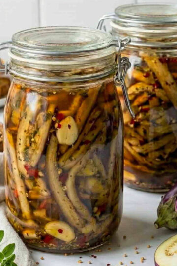 Close-up of two jars of pickled eggplant with garlic, herbs, and chili flakes, with fresh eggplant and seasonings arranged on a countertop.