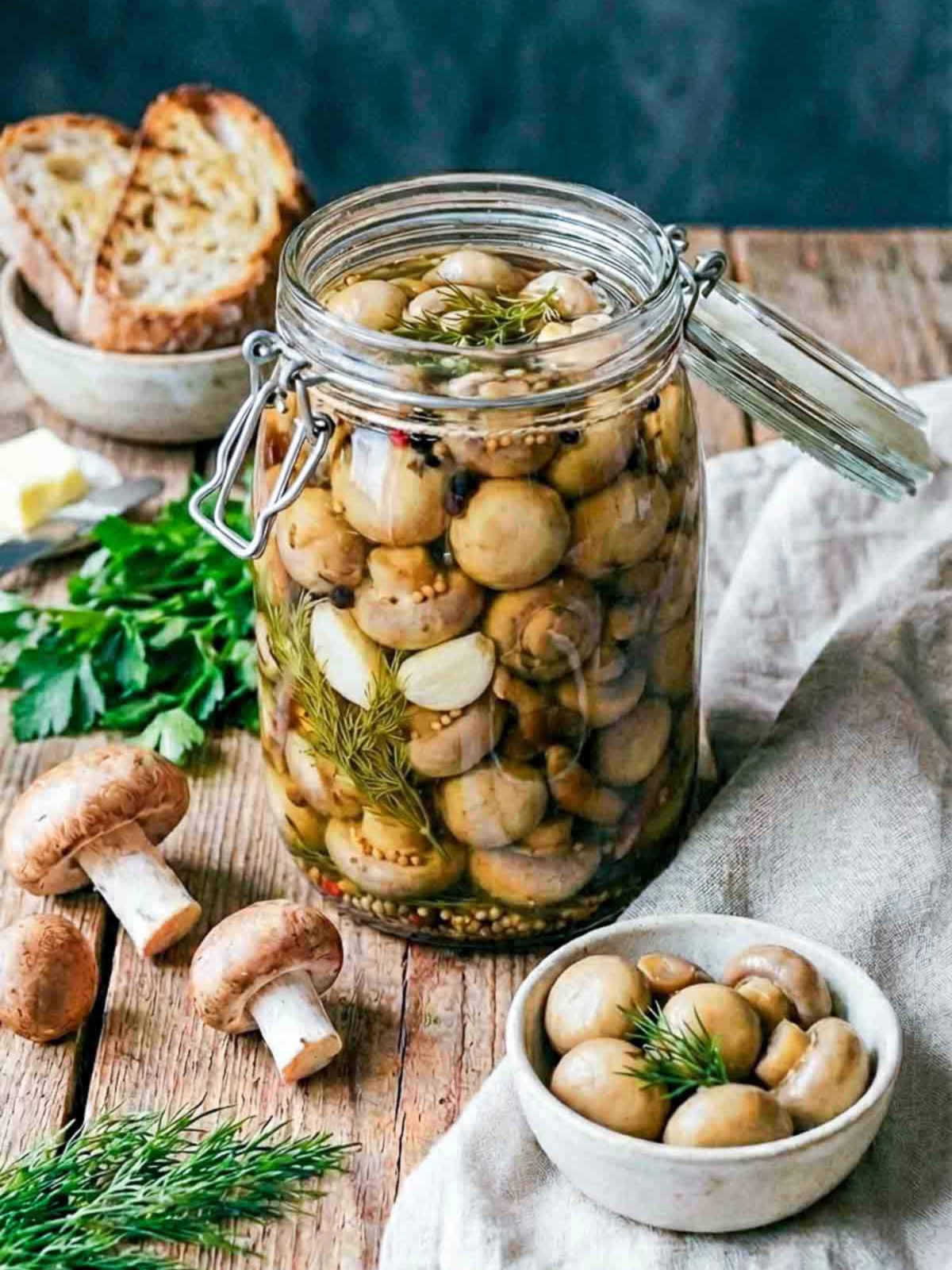 Glass jar filled with pickled mushrooms in a herb and spice brine, surrounded by fresh mushrooms, dill, parsley, and rustic bread on a wooden surface.