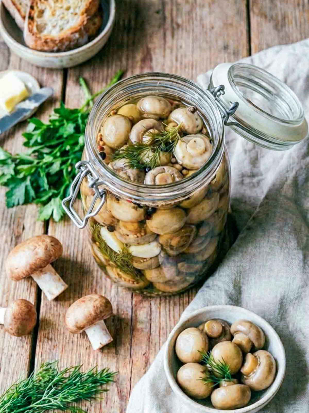 Open glass jar filled with pickled mushrooms in a herb-infused brine, topped with dill and spices, surrounded by fresh mushrooms, parsley, and rustic bread on a wooden table.