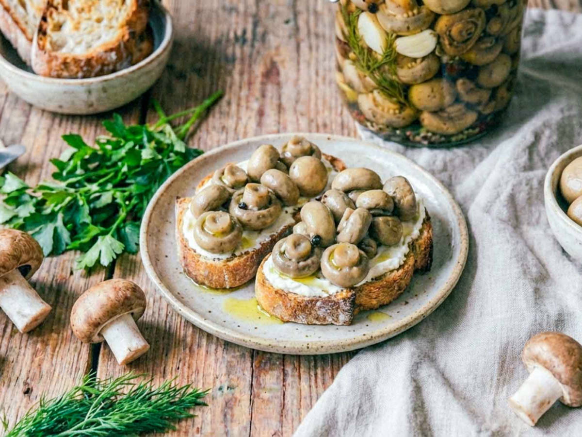 Two slices of toasted bread topped with pickled mushrooms and creamy spread, served on a plate, with a jar of pickled mushrooms, fresh herbs, and whole mushrooms on a rustic wooden table.