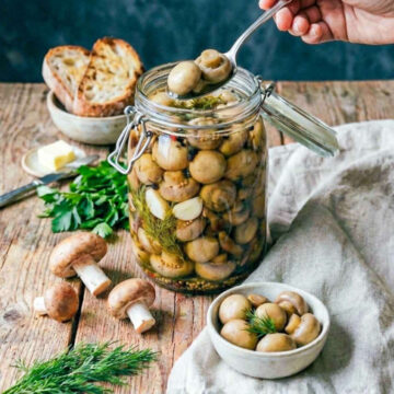 Hand lifting pickled mushrooms with a spoon from a glass jar filled with herbs and spices, surrounded by fresh mushrooms, bread, and greens on a rustic wooden table.