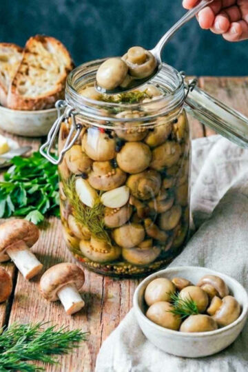 Hand lifting pickled mushrooms with a spoon from a glass jar filled with herbs and spices, surrounded by fresh mushrooms, bread, and greens on a rustic wooden table.