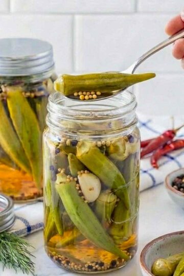 Close up of a hand holding pickled okra using a spoon over the jar.
