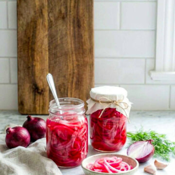 Jar of pickled red onions on a kitchen counter with fresh onions, garlic, and herbs nearby.