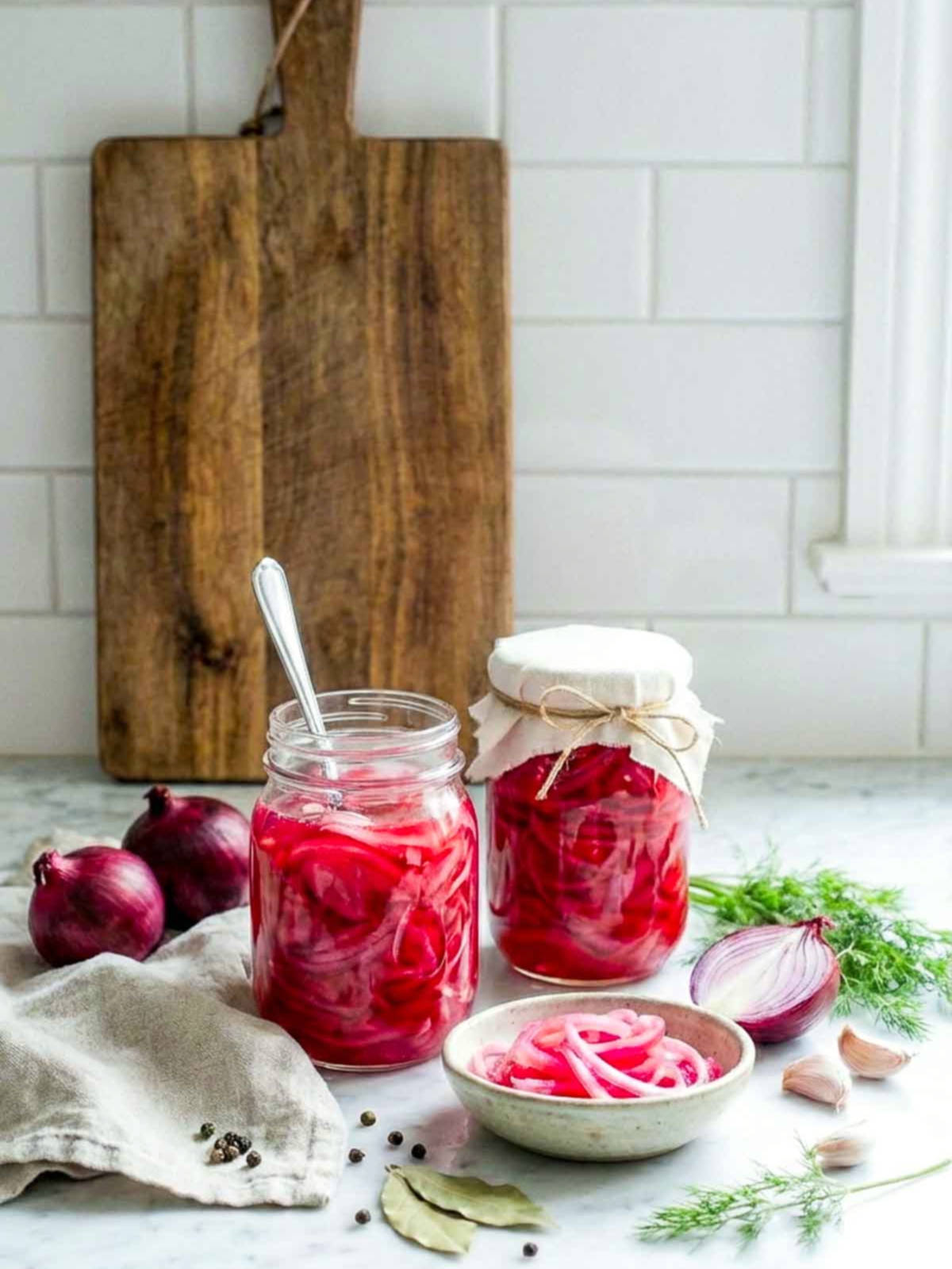 Jar of pickled red onions on a kitchen counter with fresh onions, garlic, and herbs nearby.