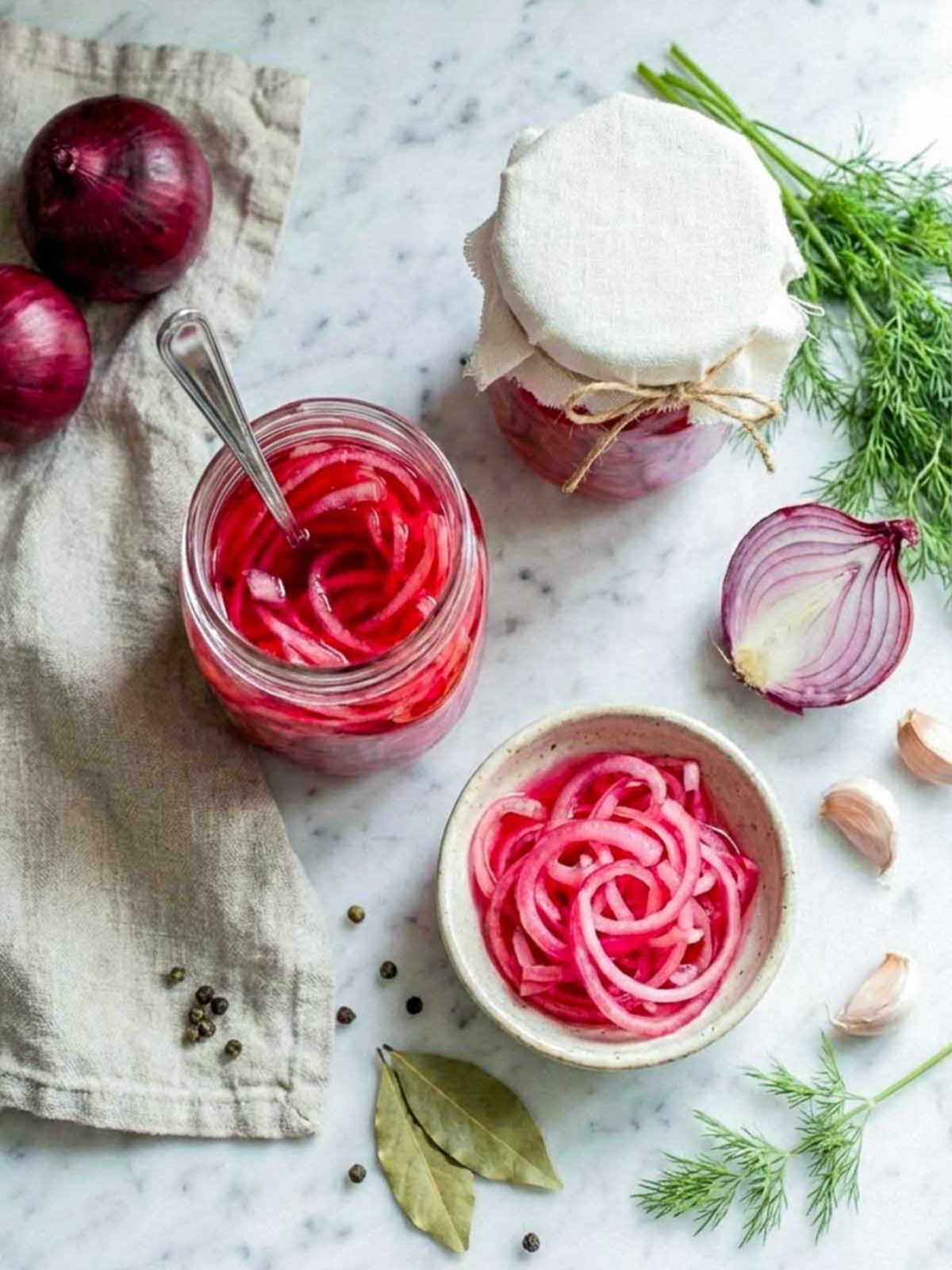 Jar and bowl of pickled red onions with fresh onions, garlic, dill, and spices on a marble surface.