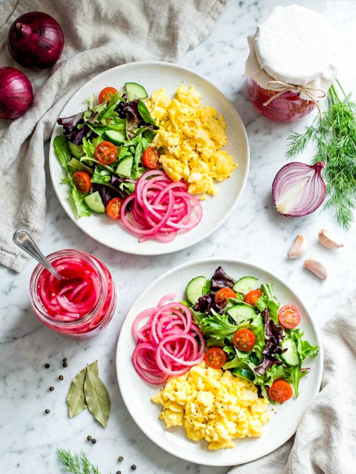 Two plates with scrambled eggs, fresh salad, and pickled red onions beside jars of onions and herbs.