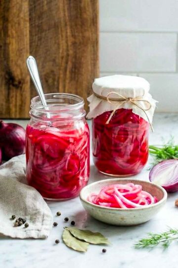 Jars of pickled red onions with fresh onions, garlic, dill, and spices on a kitchen counter.