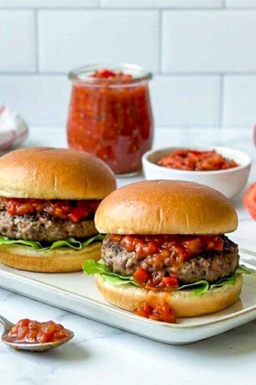 Two hamburgers topped with red pepper relish on a plate, with a jar of relish and fresh tomatoes in the background.