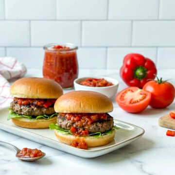 Two burgers topped with red hamburger relish on a serving plate, with a jar of relish, tomatoes, and bell peppers in the background.