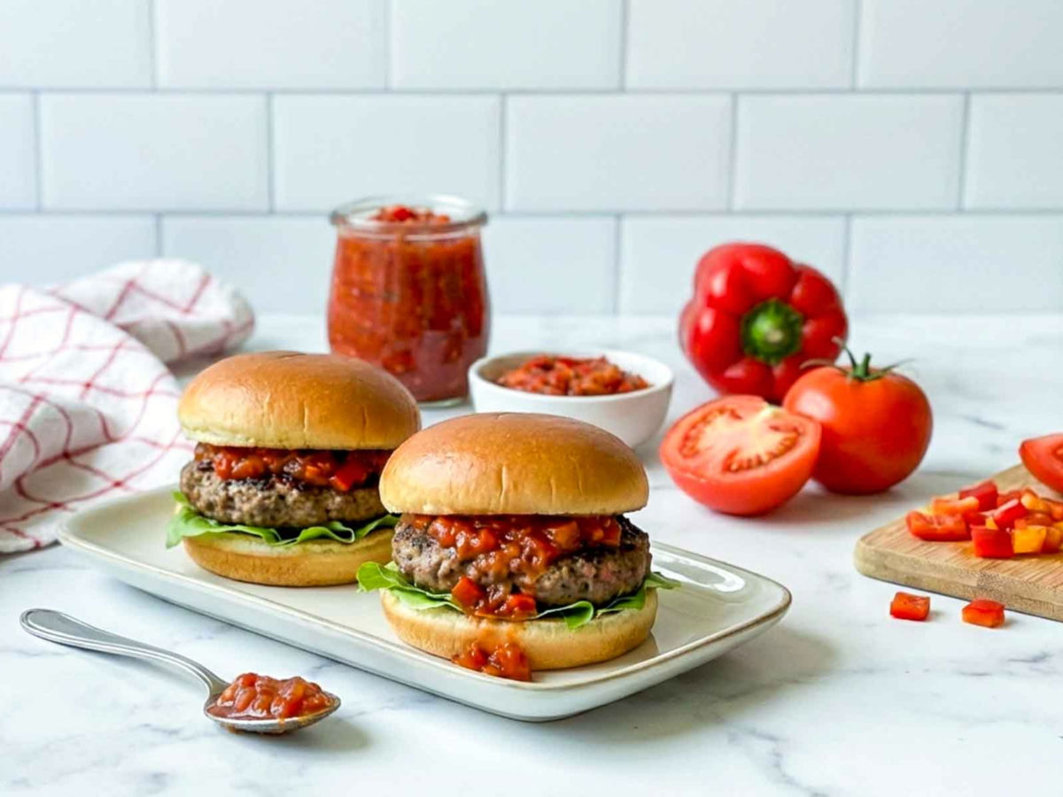 Two burgers topped with red hamburger relish on a serving plate, with a jar of relish, tomatoes, and bell peppers in the background.
