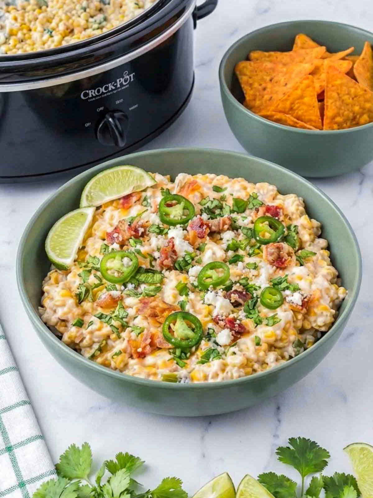 Roasted corn jalapeño street corn dip topped with bacon, cheese, cilantro, and lime wedges, served with tortilla chips and a crockpot in the background.