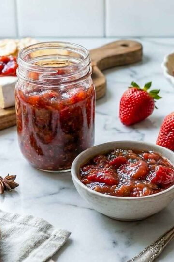 Jar and bowl of strawberry chutney with toast, samosas, fresh strawberries, and star anise in the background.