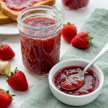 Jar of strawberry jam with a spoonful in a bowl, surrounded by fresh strawberries and ginger.