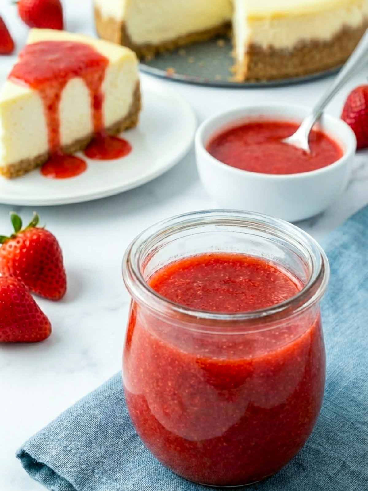 Jar of strawberry sauce with a bowl of sauce and a slice of cheesecake topped with strawberry sauce, surrounded by fresh strawberries.