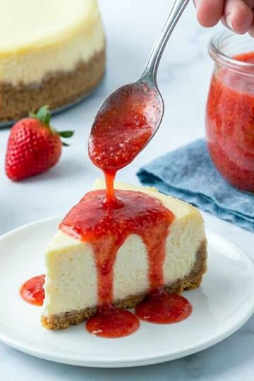 Hand pouring strawberry sauce over a slice of cheesecake, with a jar of sauce and fresh strawberries in the background.