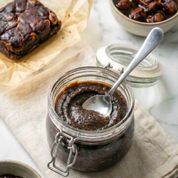Glass jar of dark tamarind paste with a spoon, beside tamarind pulp and small bowls on a cloth.
