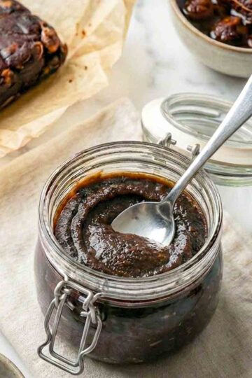 Glass jar of dark tamarind paste with a spoon, beside tamarind pulp and small bowls on a cloth.