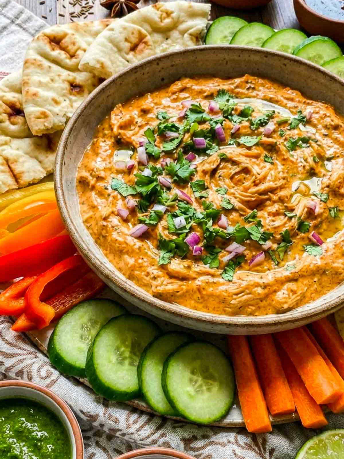Creamy tandoori chicken dip garnished with cilantro and diced red onion, served with naan, cucumber slices, carrot sticks, and bell pepper strips.