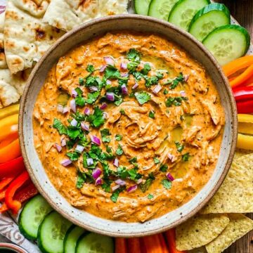 Top view of creamy tandoori chicken dip garnished with cilantro and diced red onion, surrounded by naan, tortilla chips, cucumbers, and sliced peppers.