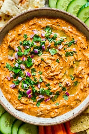 Top view of creamy tandoori chicken dip garnished with cilantro and diced red onion, surrounded by naan, tortilla chips, cucumbers, and sliced peppers.