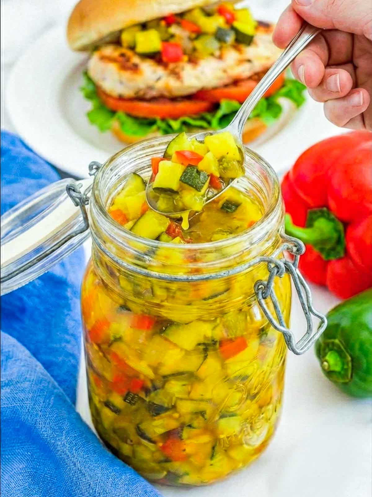 Spoon scooping homemade zucchini relish with diced zucchini and peppers from a glass jar, with a chicken sandwich in the background.