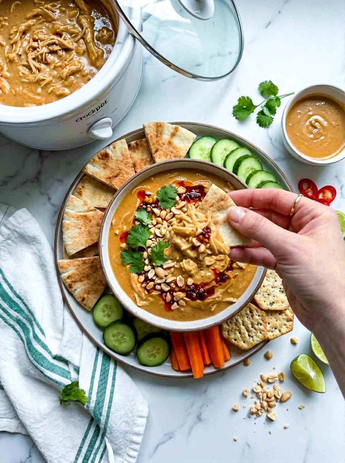 Hand dipping a pita wedge into the peanut chicken dip, bowl surrounded by crackers, carrot sticks and cucumber slices.