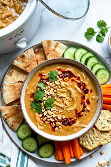 Peanut chicken dip served with pita bread, cucumber, carrots, crackers, and peanuts on top.