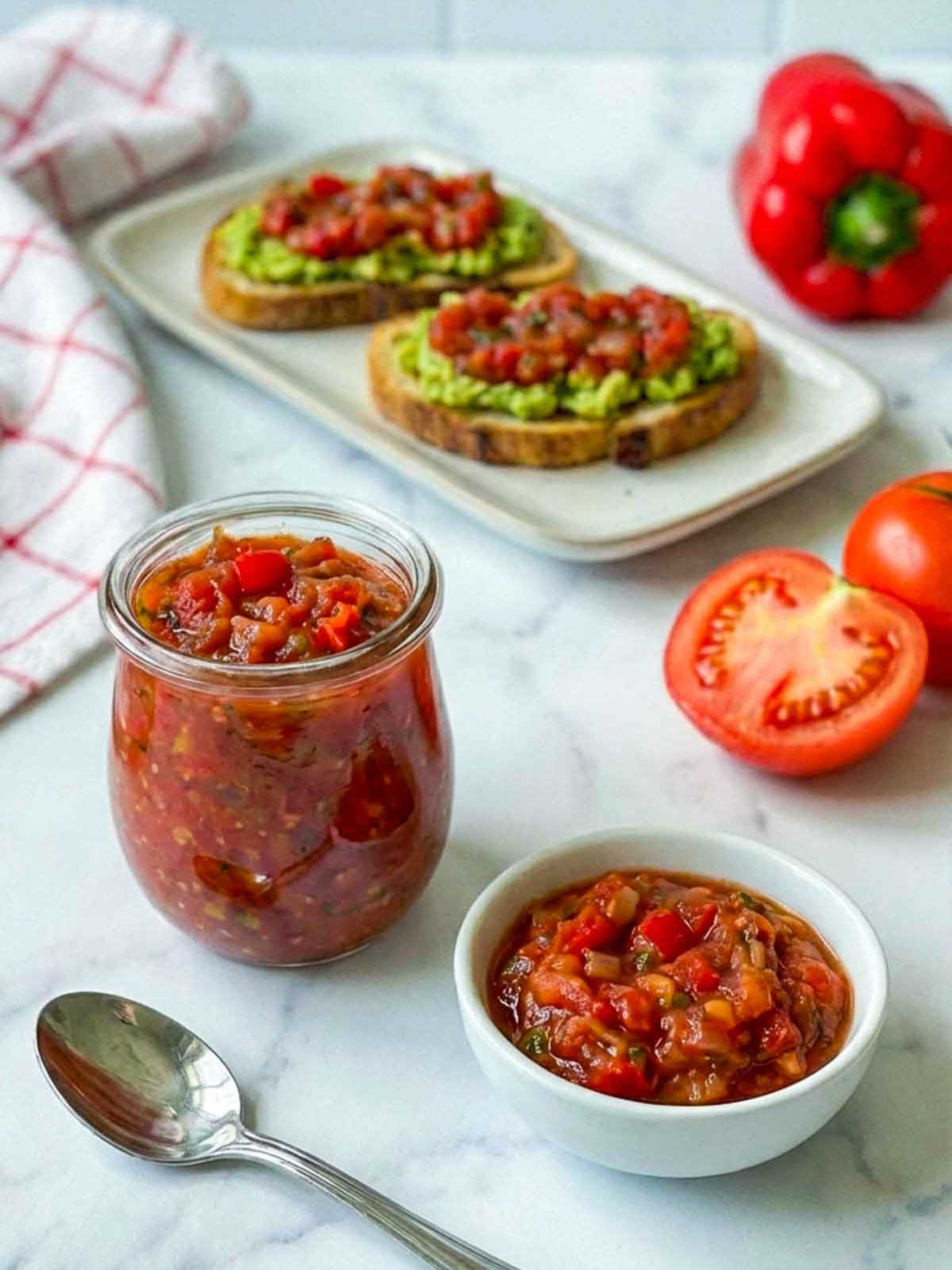 Tomato relish in jar with avocado toast and fresh tomatoes on marble surface.