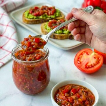 Hand scooping chunky tomato relish from jar with avocado toast in background