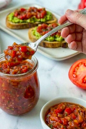 Hand scooping chunky tomato relish from jar with avocado toast in background