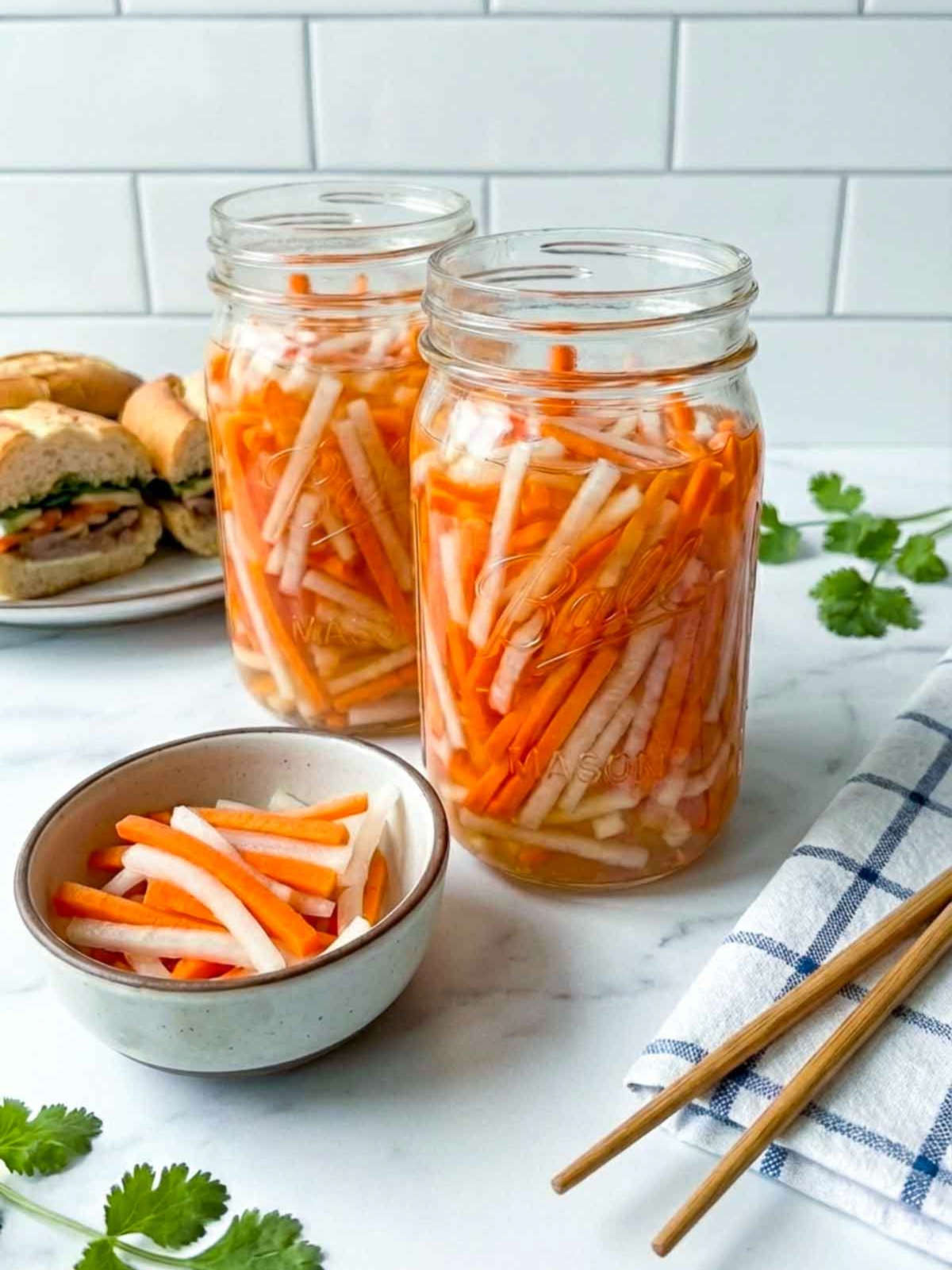 Two jars of pickled carrots and daikon (do chua) on a kitchen counter, with a small bowl of the pickles, chopsticks, and a sandwich in the background.