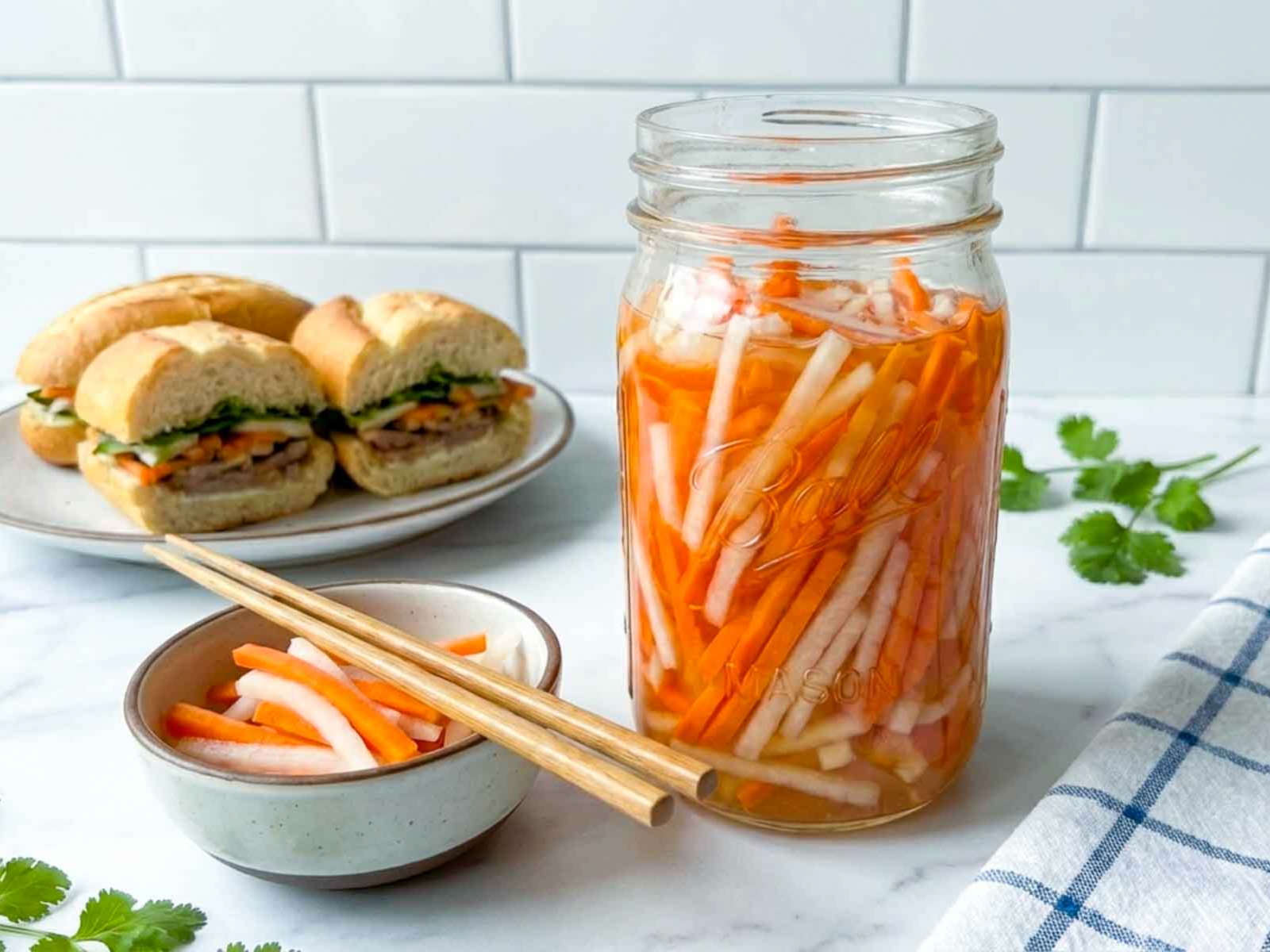 Jar of pickled carrots and daikon with a small bowl and chopsticks in front, and banh mi sandwiches in the background on a kitchen counter.