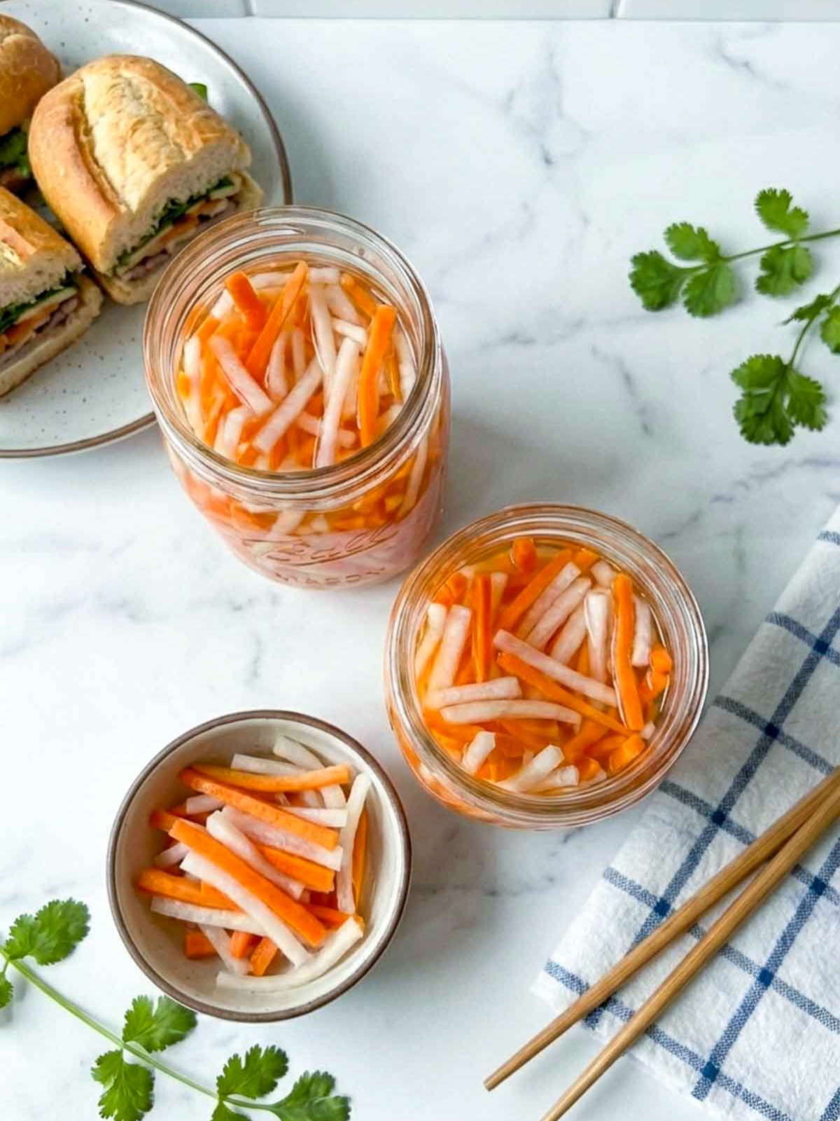 Top view of two jars of Vietnamese pickled carrots and daikon on a kitchen counter with a small bowl, chopsticks, fresh cilantro, and banh mi sandwiches in the background.
