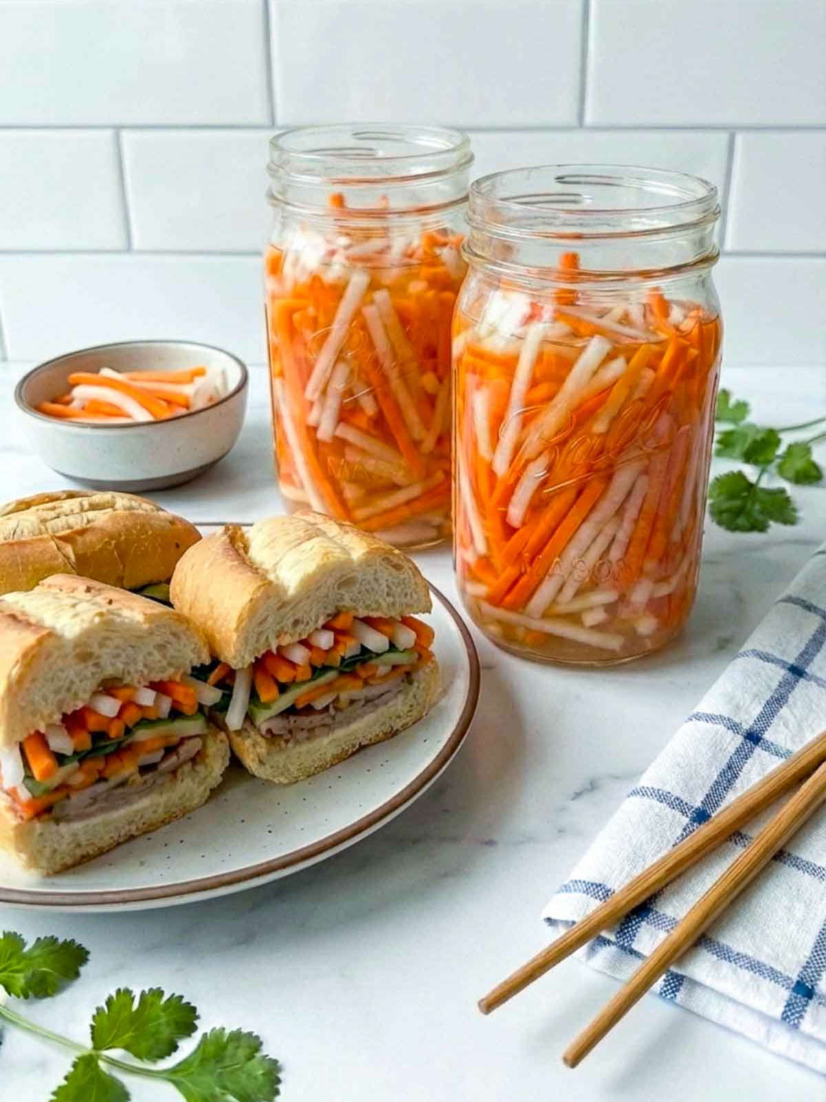 Plate of banh mi sandwiches with pickled carrots and daikon, alongside two jars of the pickles, chopsticks, and fresh cilantro on a kitchen counter.