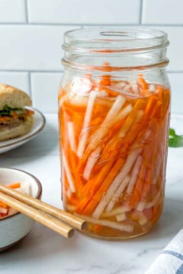 Jar of Vietnamese pickled carrots and daikon on a kitchen counter with a small bowl, chopsticks, fresh cilantro, and banh mi sandwiches in the background.