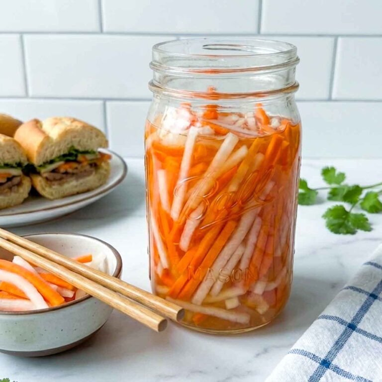 Jar of Vietnamese pickled carrots and daikon on a kitchen counter with a small bowl, chopsticks, fresh cilantro, and banh mi sandwiches in the background.