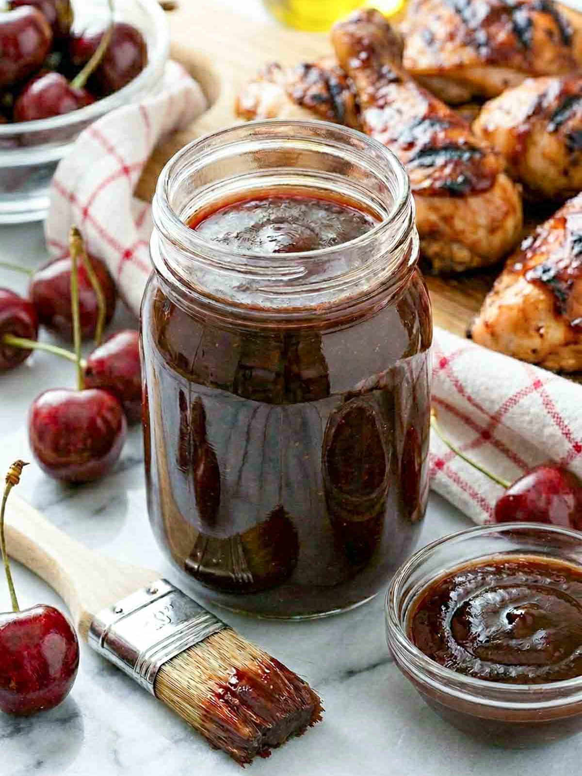 A mason jar filled with deep mahogany cherry BBQ sauce on a white marble surface, with a basting brush and small glass bowl of sauce in the foreground, fresh cherries scattered around, and glazed grilled chicken pieces on a wooden board in the background.