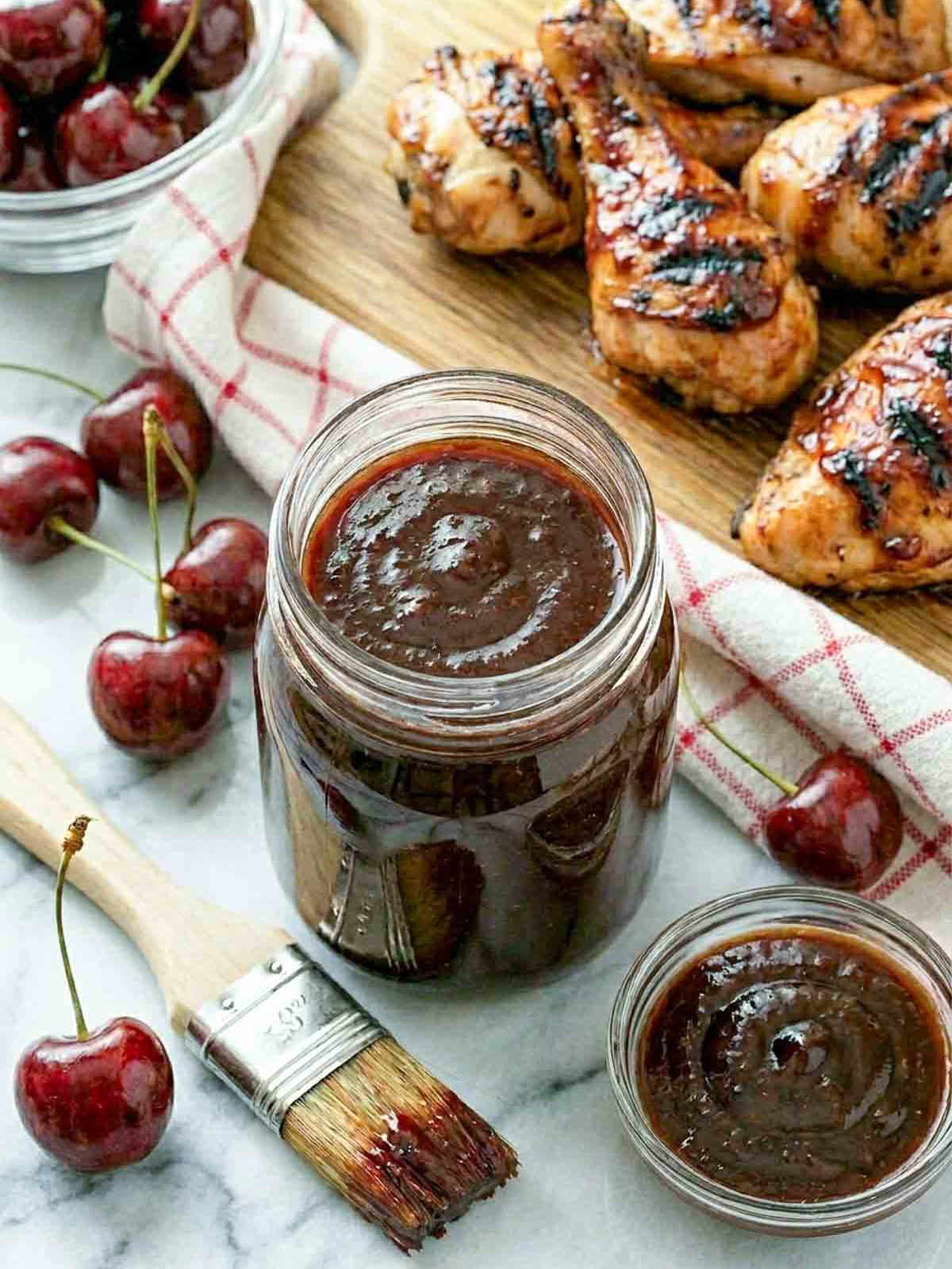 Overhead-angle view of a mason jar of thick cherry BBQ sauce with a rich, glossy surface, surrounded by fresh cherries and a basting brush on white marble, grilled chicken thighs on a wooden board with a red-checked cloth behind.