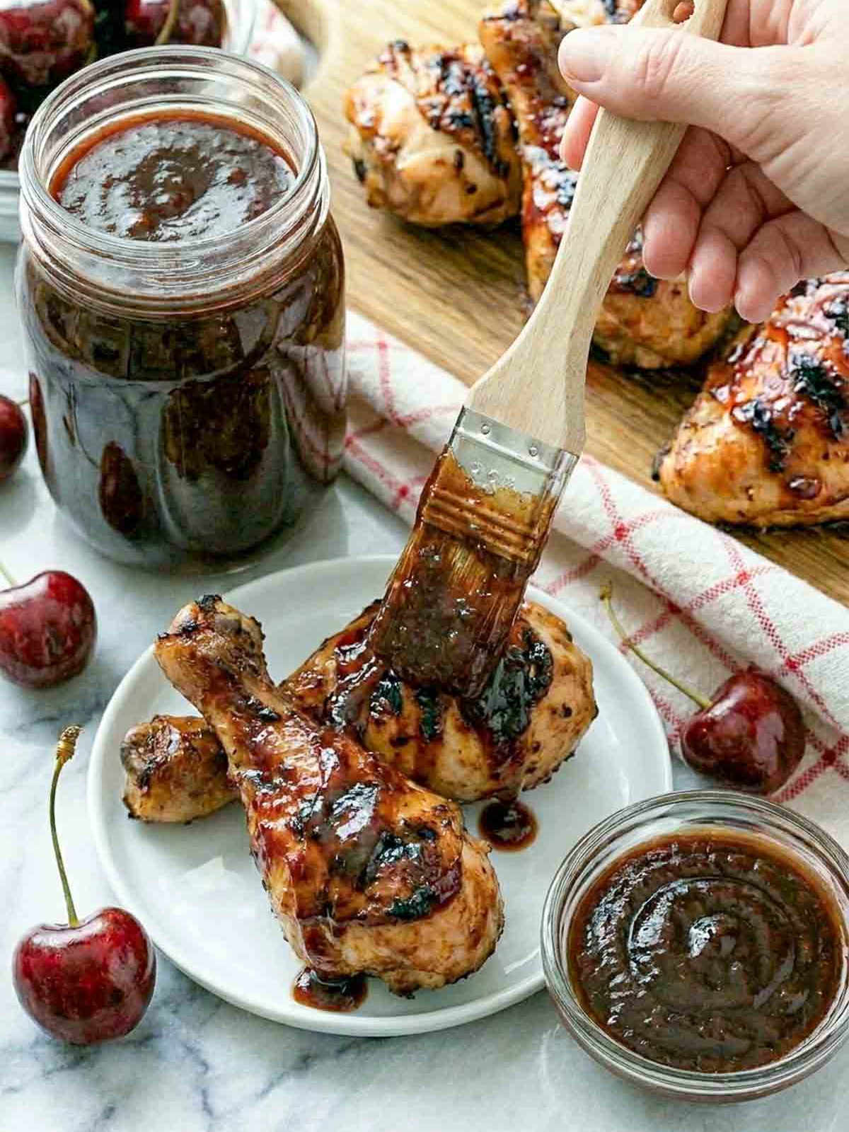 A hand brushing cherry BBQ sauce onto two grilled chicken drumsticks on a small white plate, with a mason jar of sauce, small glass bowl, and additional glazed chicken on a wooden board arranged on white marble.