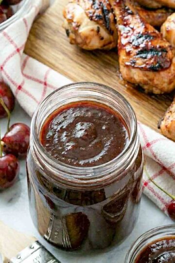 Square-cropped close-up of a mason jar of deep cherry BBQ sauce centered on white marble, flanked by a basting brush and small bowl of sauce, with fresh cherries and glazed grilled chicken in the background.