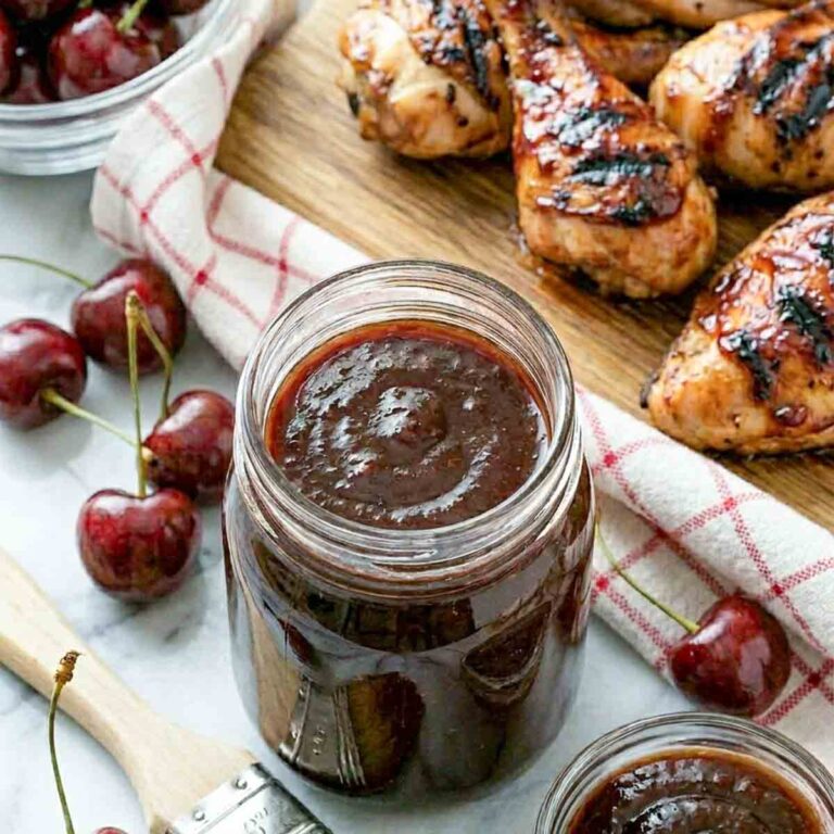 Square-cropped close-up of a mason jar of deep cherry BBQ sauce centered on white marble, flanked by a basting brush and small bowl of sauce, with fresh cherries and glazed grilled chicken in the background.