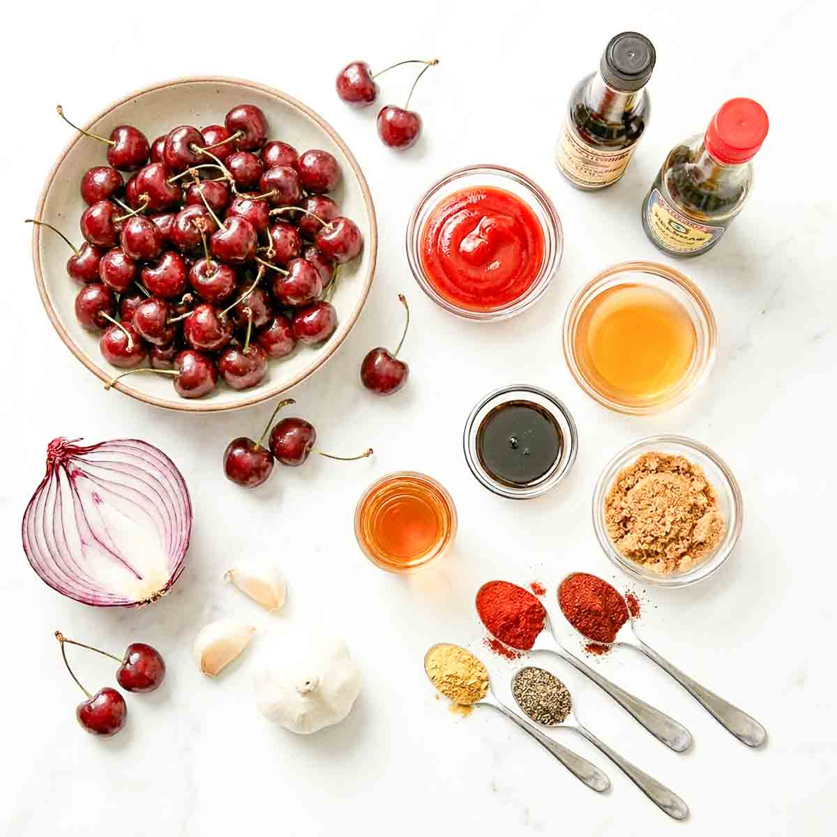 Flatlay of cherry BBQ sauce ingredients on a white marble surface: a large bowl of fresh cherries, halved red onion, whole garlic bulb and loose cloves, small bowls of ketchup, apple cider vinegar, balsamic vinegar, honey, and brown sugar, with Worcestershire sauce bottle and spoons holding smoked paprika, cayenne, black pepper, and mustard powder.